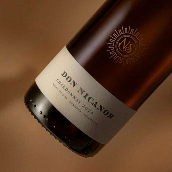Don Nicanor Varietals demonstrates heritage-forward premiumization through dual embellishment strategy: copper silkscreen symbol applied directly to glass materializes the winery's architectural legacy, while cotton substrate with dry-stamped logo creates tactile authenticity that signals artisanal craftsmanship to quality-conscious wine consumers. Design by Tridimage